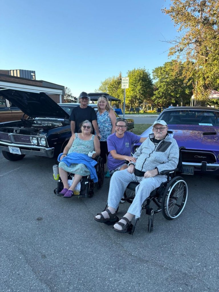 Residents and family gathered between a Beaumont and a purple Dodge Challenger