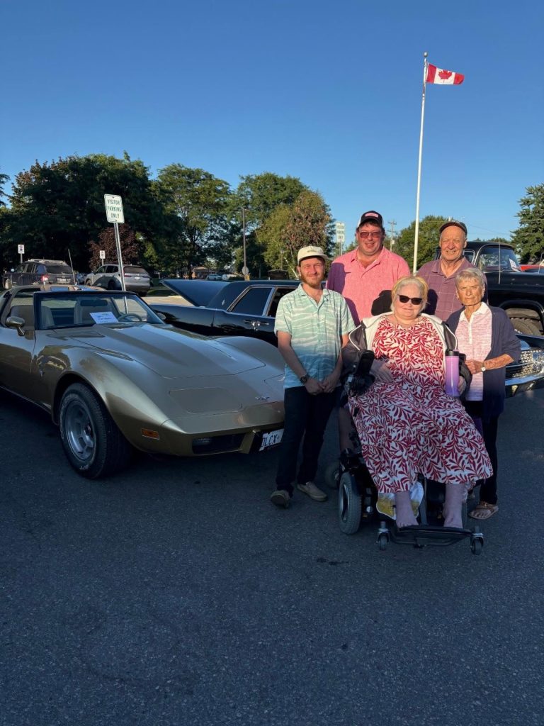 Residents and visitors beside a gold Corvette, with the Canadian flag flying behind